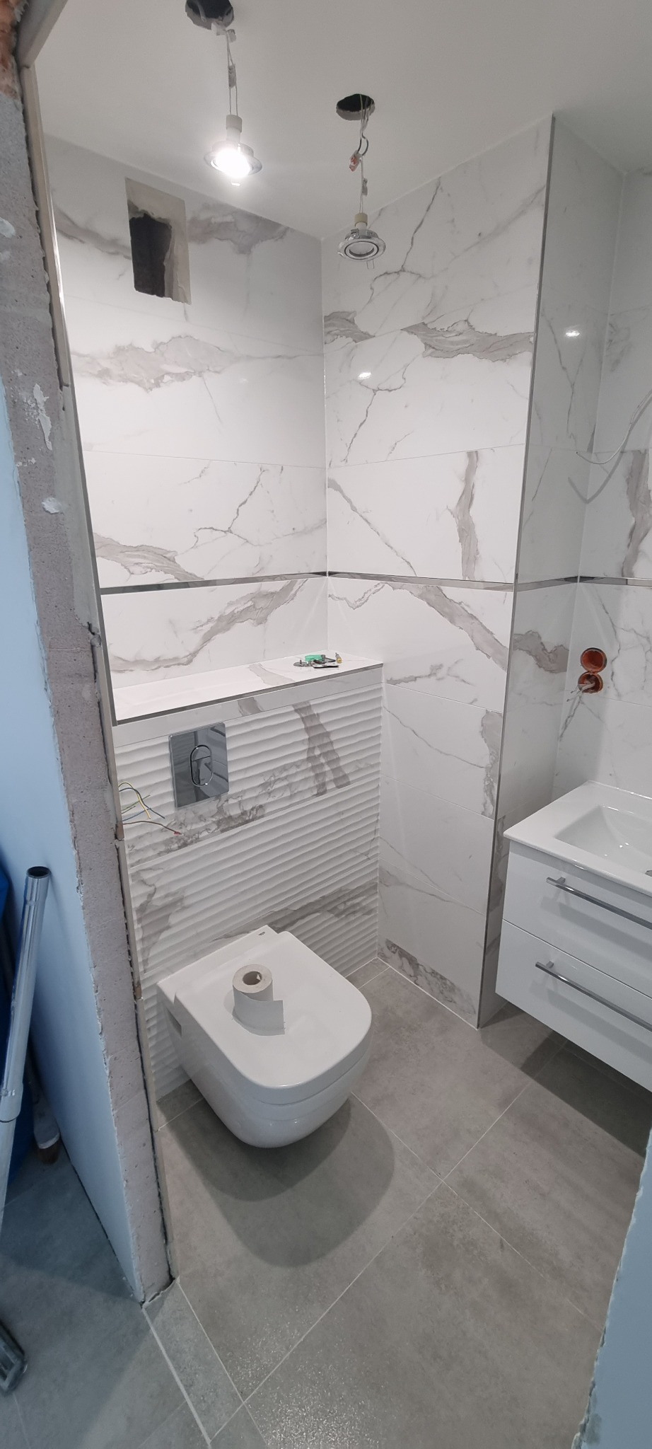 Wnętrze łazienki w trakcie remontu: częściowo ułożone płytki imitujące marmur, podwieszana toaleta z rolką papieru, niedokończone obramowanie drzwi, widoczne instalacje elektryczne.