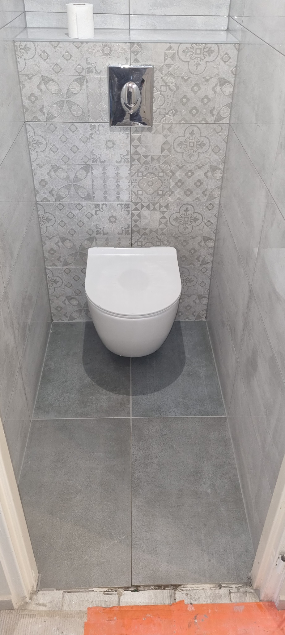 Podwieszana toaleta w szarej łazience z dekoracyjnymi płytkami na ścianie, widoczny fragment podłogi z pomarańczową folią zabezpieczającą.