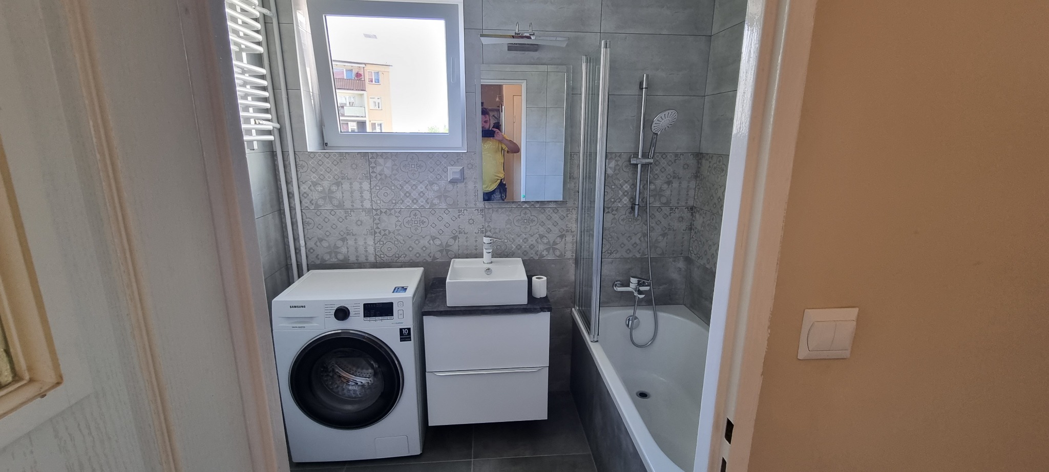 Wyremontowana łazienka z szarymi płytkami, białą pralką Samsung, umywalką na szafce i wanną z parawanem, widok zza otwartych drzwi.