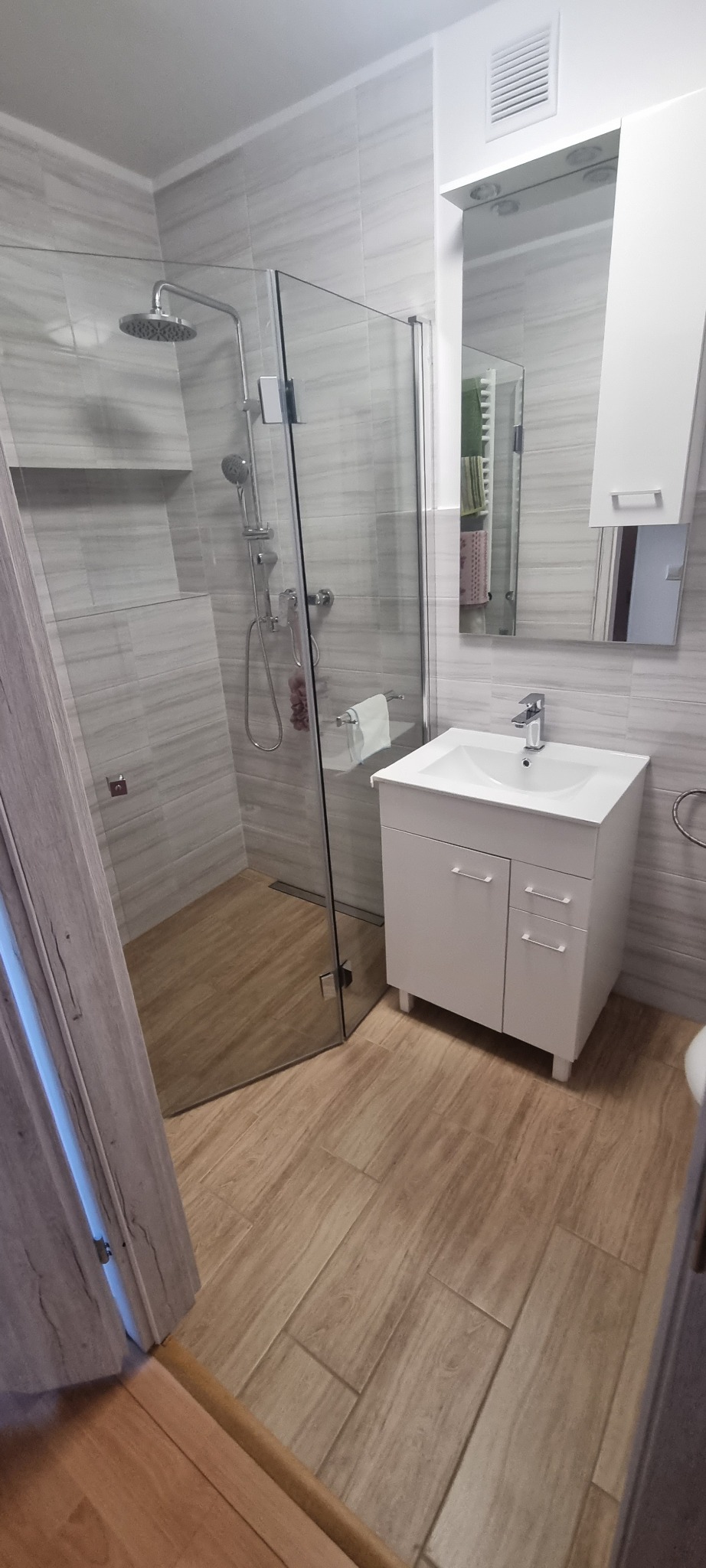 Nowoczesna łazienka po renowacji w Pyrzycach: kabina prysznicowa typu walk-in z deszczownicą, umywalka z szafką i lustro z oświetleniem, wykończona płytkami imitującymi drewno.