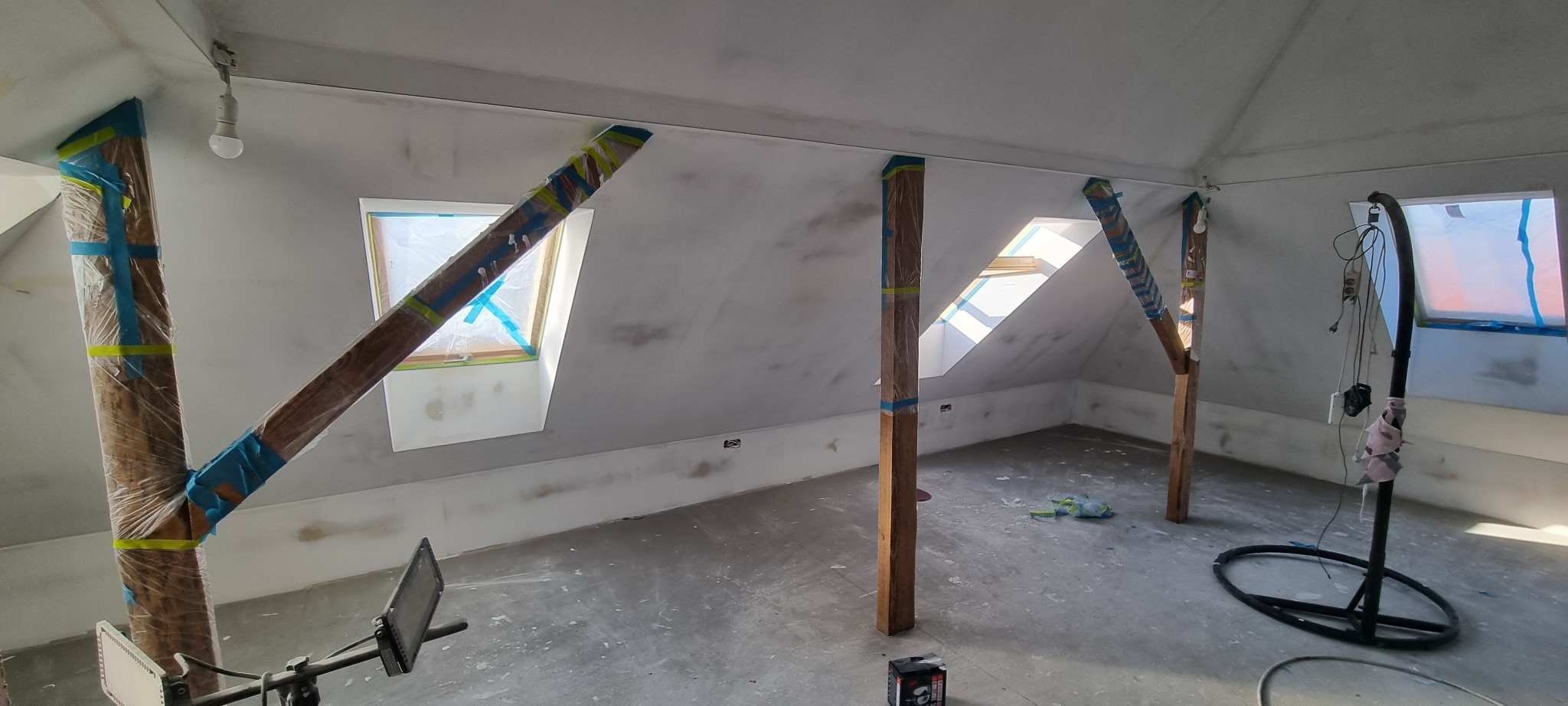 Poddasze w trakcie szpachlowania, drewniane słupy zabezpieczone folią i taśmą malarską, okna dachowe zakryte folią, podłoga pokryta szpachlą, widoczne lampy budowlane i kable.
