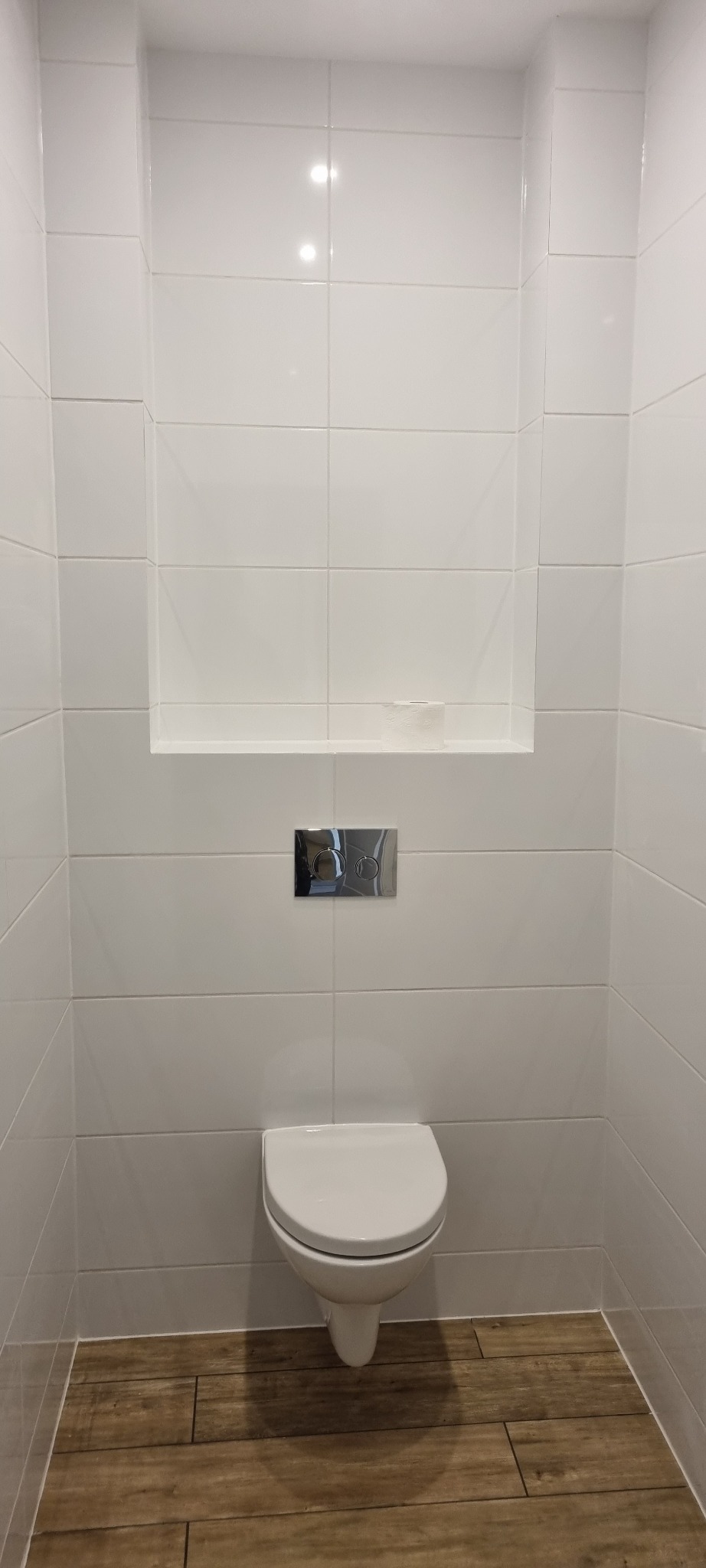 Nowoczesna toaleta z białymi, błyszczącymi płytkami i drewnopodobną podłogą, wnęka z rolką papieru toaletowego nad spłuczką.