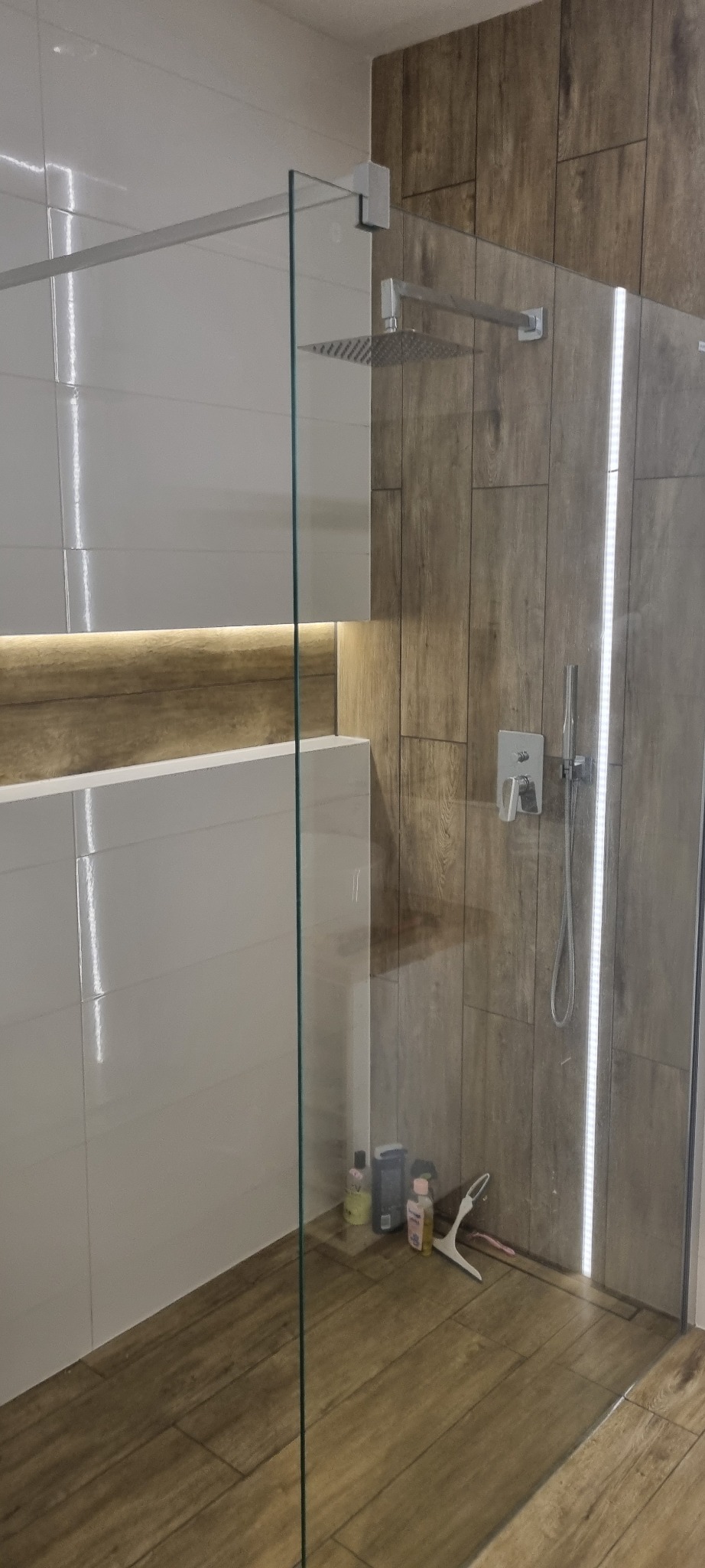 Nowoczesna kabina prysznicowa typu walk-in z deszczownicą, wykończona białymi i drewnopodobnymi płytkami, z podświetleniem LED i widocznymi akcesoriami kosmetycznymi na podłodze.
