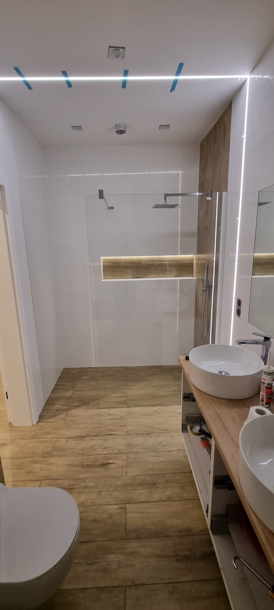 Nowoczesna łazienka z kabiną prysznicową typu walk-in, drewnopodobną podłogą, białymi płytkami ściennymi i podwójną umywalką nablatową na drewnianym blacie; widoczne niebieskie taśmy malarskie...
