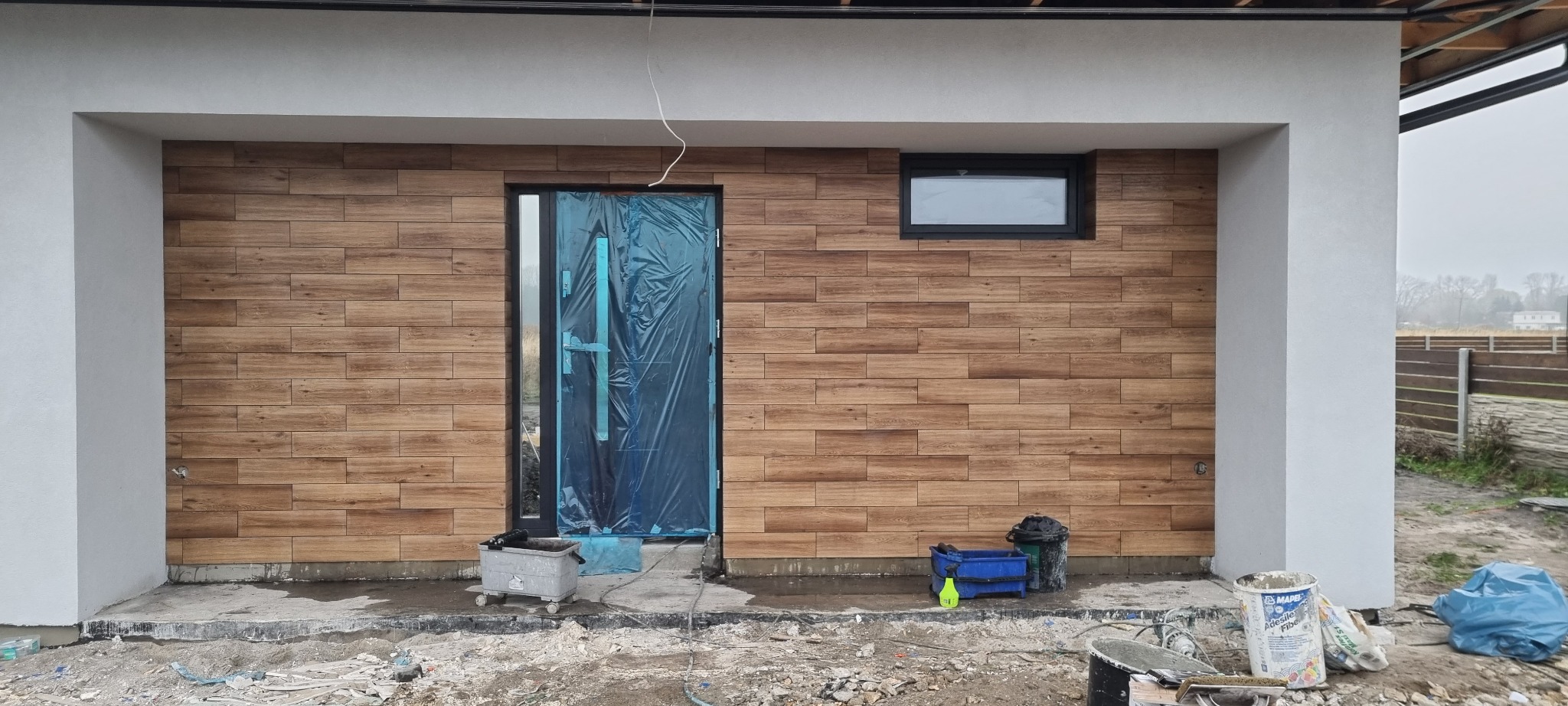 Elewacja budynku wykończona płytkami imitującymi drewno, z oknem i drzwiami zabezpieczonymi folią, widoczne narzędzia i materiały budowlane na ziemi.