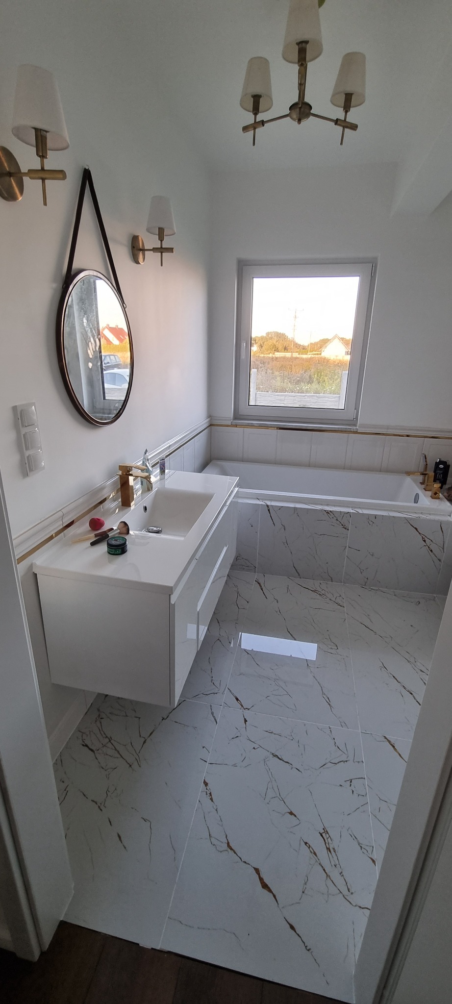 Nowoczesna łazienka z białymi płytkami imitującymi marmur, wisząca szafka pod umywalką z złotą armaturą, okrągłe lustro na skórzanym pasku i złote oświetlenie.