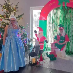 Kobiety w kostiumach: królowa śniegu w błękitnej sukni i elf w zielono-czerwonym stroju, dekoracje świąteczne, choinka i dmuchany łuk 'Merry Christmas'.