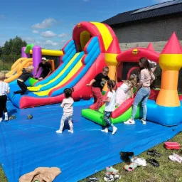 Dzieci bawią się na kolorowym, dmuchanym zamku z dwiema zjeżdżalniami, rozłożonym na niebieskiej plandece na trawie; w tle budynek z ciemnym dachem i fotowoltaiką.