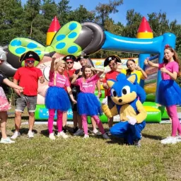 Grupa animatorów w kostiumach i przebraniach pozuje na tle dmuchanych atrakcji, w tym zjeżdżalni i zamku z motywem Myszki Miki.