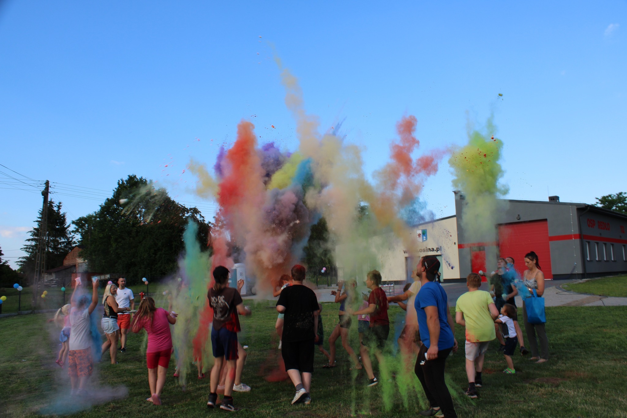 Grupa ludzi rzuca kolorowe proszki w powietrze podczas festiwalu kolorów na tle budynku OSP Redło.