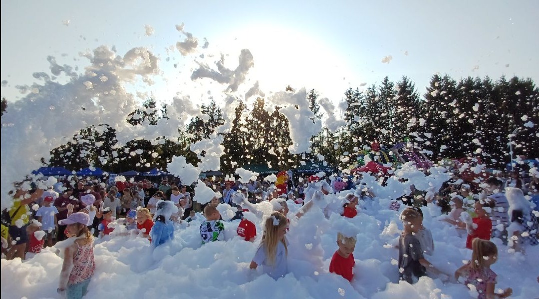 Piana party w plenerze: grupa dzieci i dorosłych bawi się w gęstej pianie pod słońcem, z widocznym tłumem w tle i zielenią drzew, tworząc radosną atmosferę.