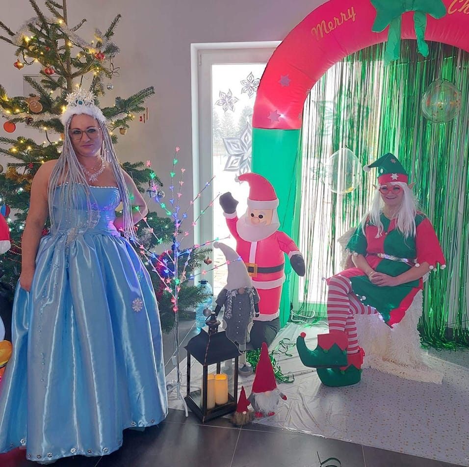 Kobiety w kostiumach: królowa śniegu w błękitnej sukni i elf w zielono-czerwonym stroju, dekoracje świąteczne, choinka i dmuchany łuk 'Merry Christmas'.