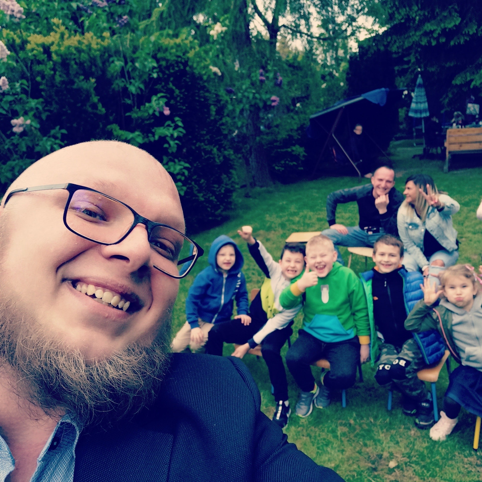 Uśmiechnięty mężczyzna w okularach robi selfie z grupą dzieci i dorosłych w ogrodzie, prawdopodobnie podczas występu lub imprezy dla dzieci.