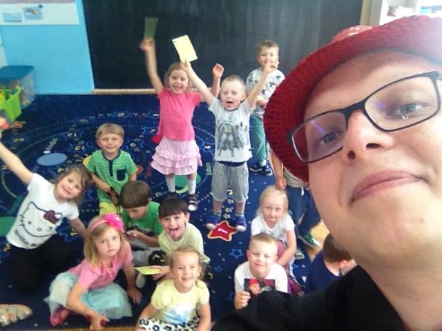 Selfie iluzjonisty w czerwonym kapeluszu i okularach na tle grupy roześmianych dzieci w przedszkolu, trzymających w rękach rekwizyty po pokazie.