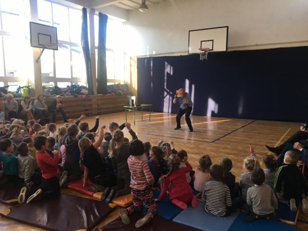 Występ dla dzieci w sali gimnastycznej: mężczyzna w koszuli i spodniach prezentuje książkę przed siedzącymi na podłodze dziećmi, na tle kosz do koszykówki i zasłoniętej ściany.