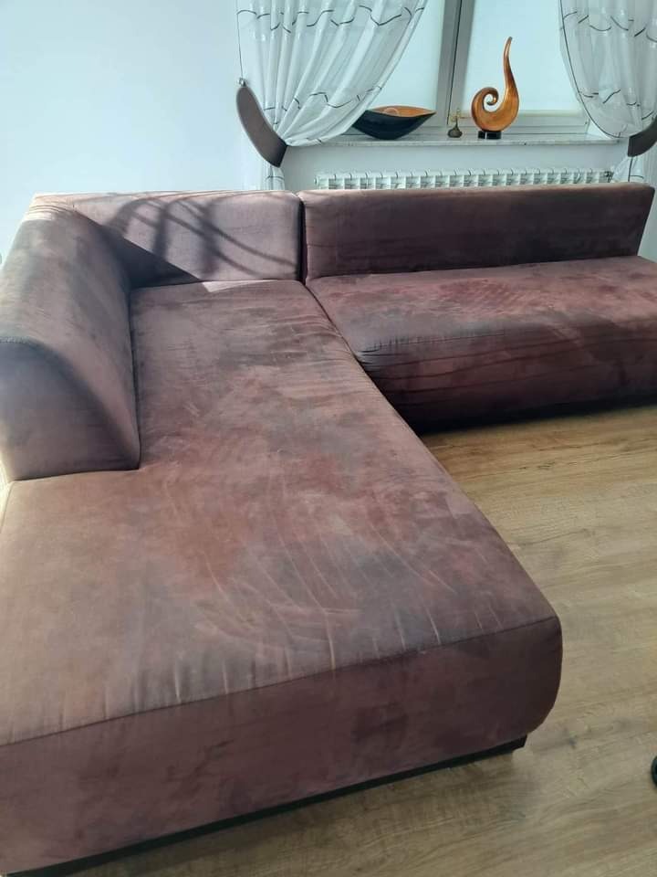 Brązowa, narożna kanapa z widocznymi śladami użytkowania i plamami na tapicerce, ustawiona w salonie z oknem w tle.