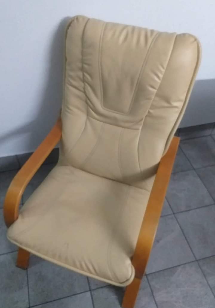 Beżowy fotel tapicerowany z drewnianymi podłokietnikami, widoczny z perspektywy góry, na tle białej ściany i szarej podłogi.