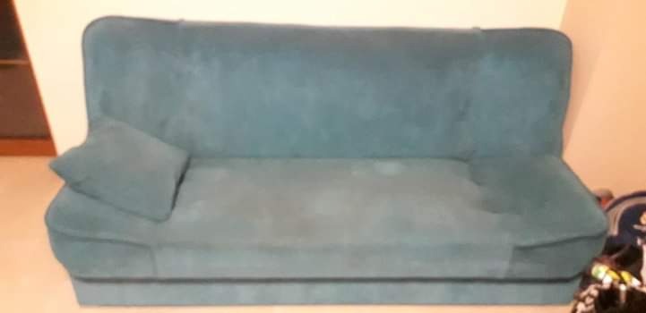 Turkusowa kanapa z poduszką w pokoju z beżowymi ścianami, widoczne ślady użytkowania.