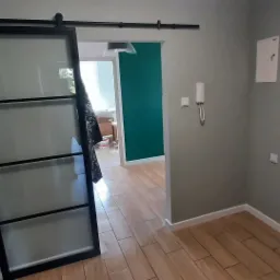 Wnętrze mieszkania w trakcie remontu: przesuwne szklane drzwi z czarną ramą, świeżo pomalowana ściana w kolorze butelkowej zieleni, drewnopodobna podłoga, widoczne zabezpieczenia folią malarską.