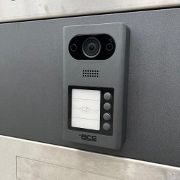 GRUPA STACHURA SP. Z O.O. - Szary wideodomofon BCS zamontowany na metalowej powierzchni. Widoczna kamera, ekran LCD i przyciski. Minimalistyczny design, nowoczesne wykończenie.
