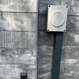 GRUPA STACHURA SP. Z O.O. - Szara skrzynka z logo Nice na słupku z szarej kostki, obok widoczny czarny przycisk. Element automatyki bramy w zbliżeniu.