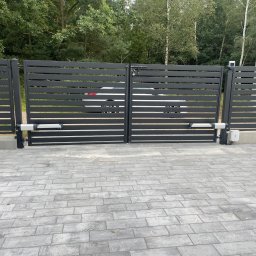 GRUPA STACHURA SP. Z O.O. - Nowoczesna, grafitowa brama przesuwna z automatyką, widoczna z zewnątrz posesji na tle zieleni. Kostka brukowa przed bramą.
