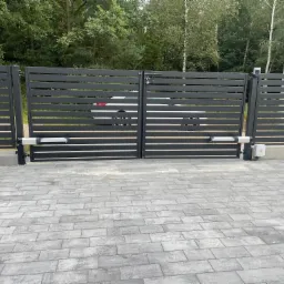 Nowoczesna, grafitowa brama przesuwna z automatyką, widoczna z zewnątrz posesji na tle zieleni. Kostka brukowa przed bramą.