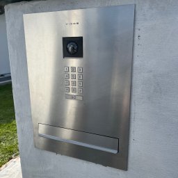 GRUPA STACHURA SP. Z O.O. - Nowoczesny, srebrny panel domofonowy Vidos z kamerą i klawiaturą numeryczną, zamontowany na szarym, surowym słupku ogrodzeniowym przy domu.