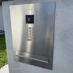 Nowoczesny, srebrny panel domofonowy Vidos z kamerą i klawiaturą numeryczną, zamontowany na szarym, surowym słupku ogrodzeniowym przy domu.