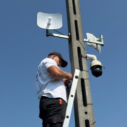 GRUPA STACHURA SP. Z O.O. - Instalator w czapce i białej koszulce, stojąc na drabinie, podłącza okablowanie do anten i czarnej kamery obrotowej zamontowanych na betonowym słupie, na tle błękitnego nieba.