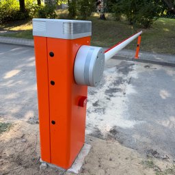 GRUPA STACHURA SP. Z O.O. - Nowoczesny, pomarańczowy szlaban automatyczny marki NICE z podniesionym ramieniem, zainstalowany na wjeździe. Widoczne czerwone światło ostrzegawcze, asfalt i zieleń w tle.