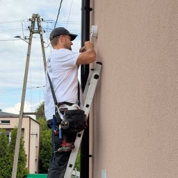 GRUPA STACHURA SP. Z O.O. - Technik na drabinie montuje biały, okrągły element na jasnobrązowej elewacji budynku, obok czarnej rynny. W tle słupy energetyczne.