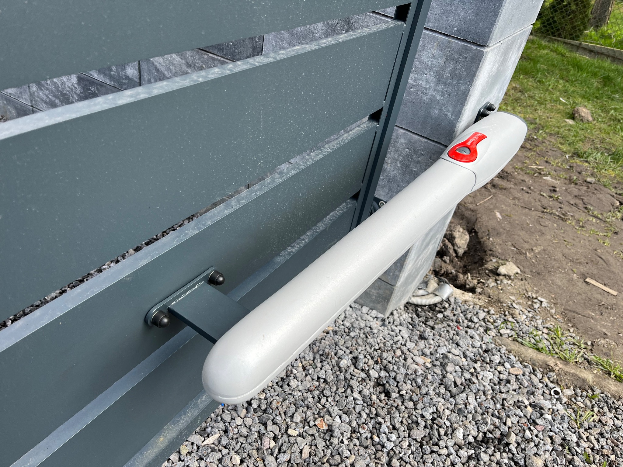 Szary automat do bramy segmentowej, zamontowany na słupku z kostki i metalowych panelach, widziany z góry na tle żwiru i trawy.