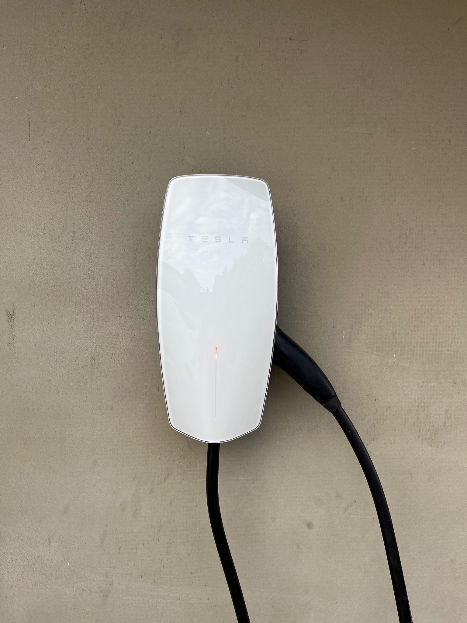 Biała ładowarka Tesla Wall Connector, zamontowana na szarej ścianie, z podłączonym czarnym kablem. Widoczna subtelna dioda LED.