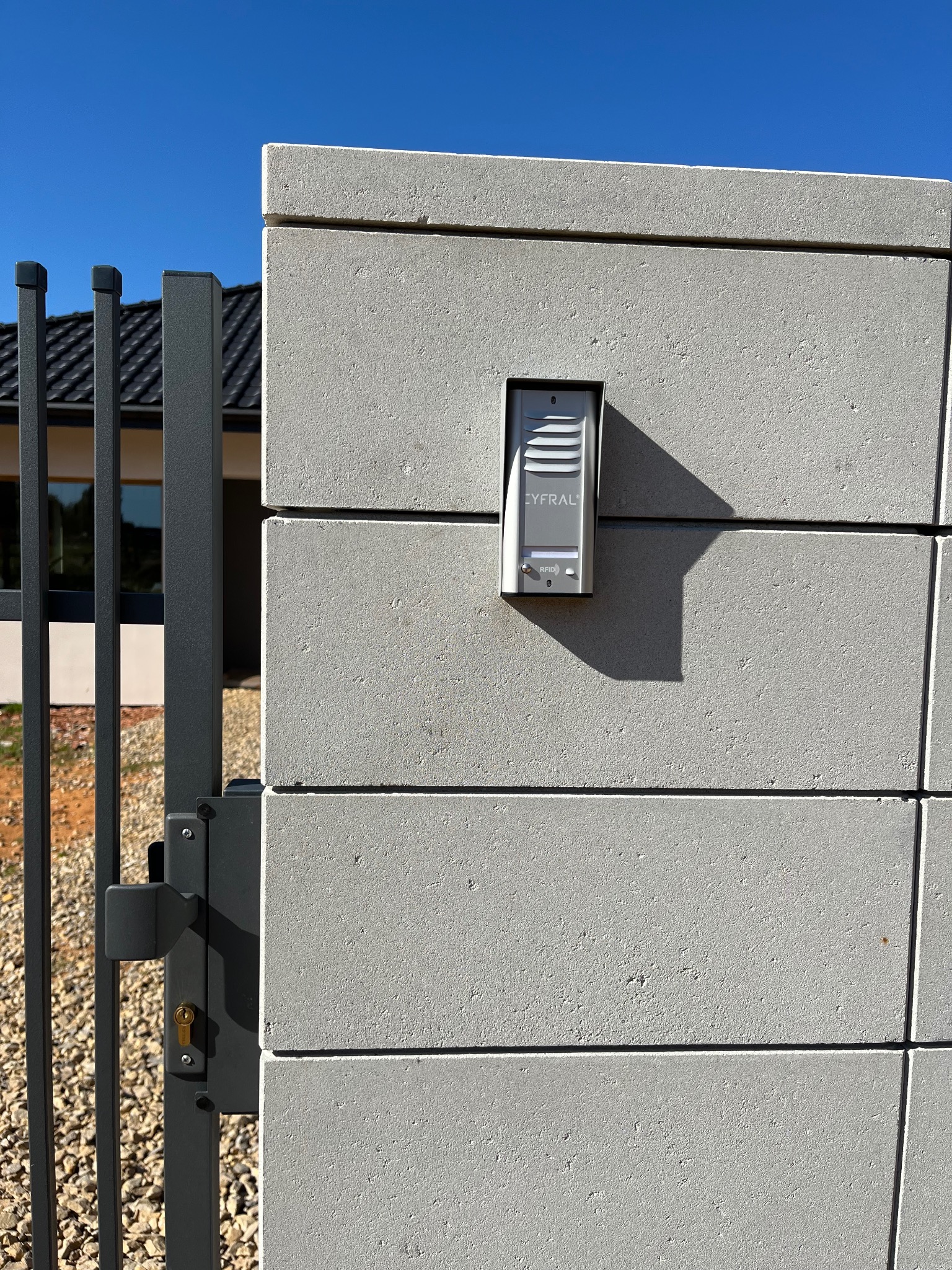 Domofon Cyfral zamontowany na betonowym słupku ogrodzenia, obok furtki. Widoczne elementy domu w tle, błękitne niebo.