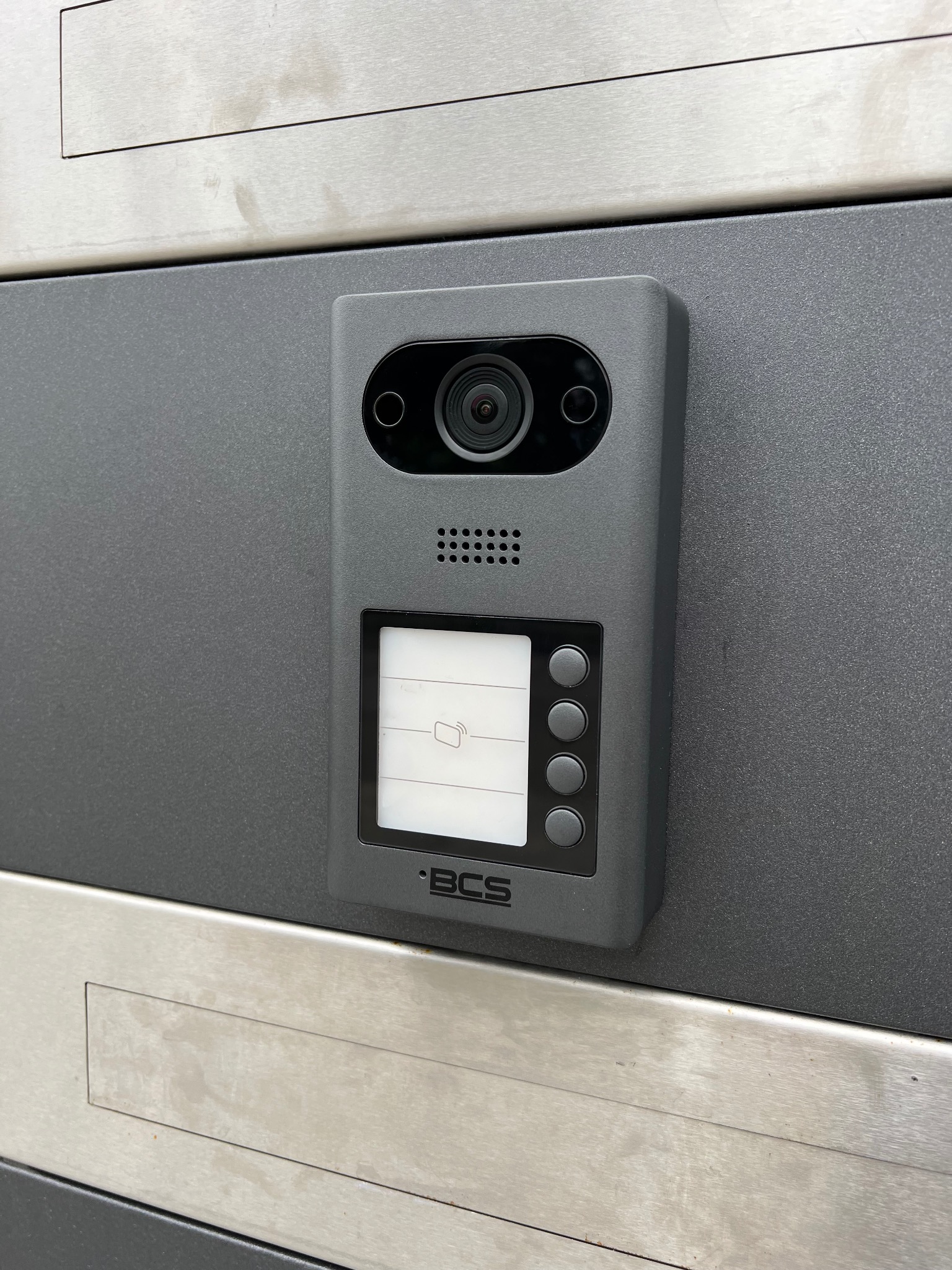 Szary wideodomofon BCS zamontowany na metalowej powierzchni. Widoczna kamera, ekran LCD i przyciski. Minimalistyczny design, nowoczesne wykończenie.