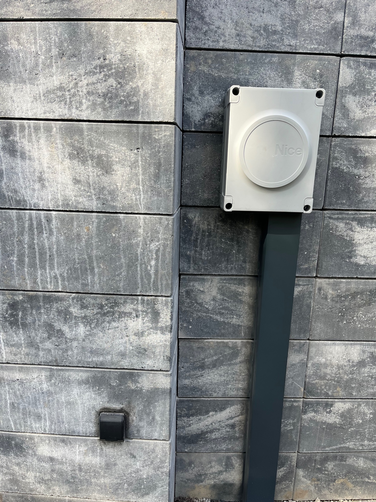 Szara skrzynka z logo Nice na słupku z szarej kostki, obok widoczny czarny przycisk. Element automatyki bramy w zbliżeniu.