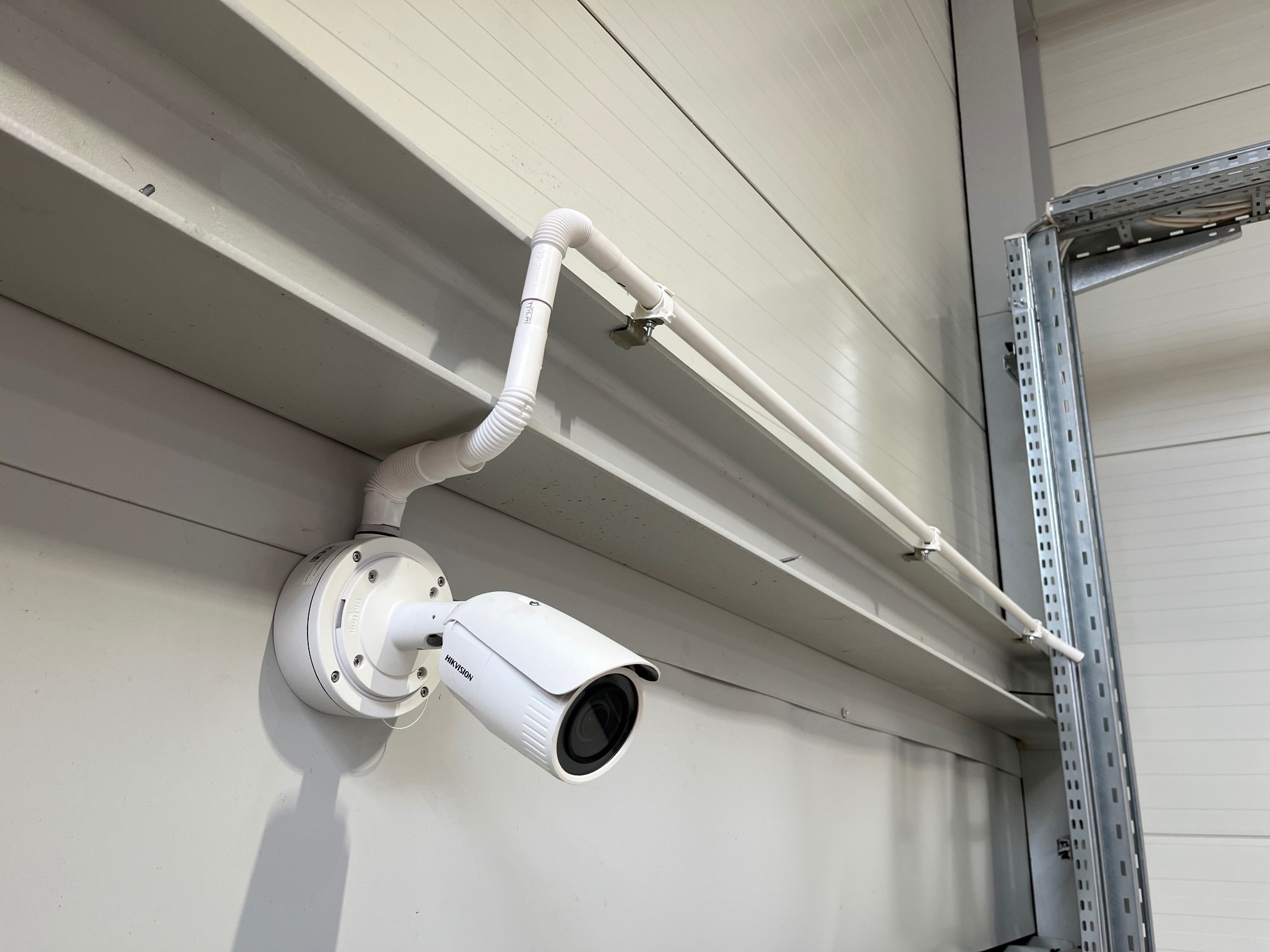 Zamontowana kamera monitoringu Hikvision na szarej ścianie z blachy trapezowej. Białe rurki osłonowe prowadzą do kamery. Widoczny fragment metalowego regału.