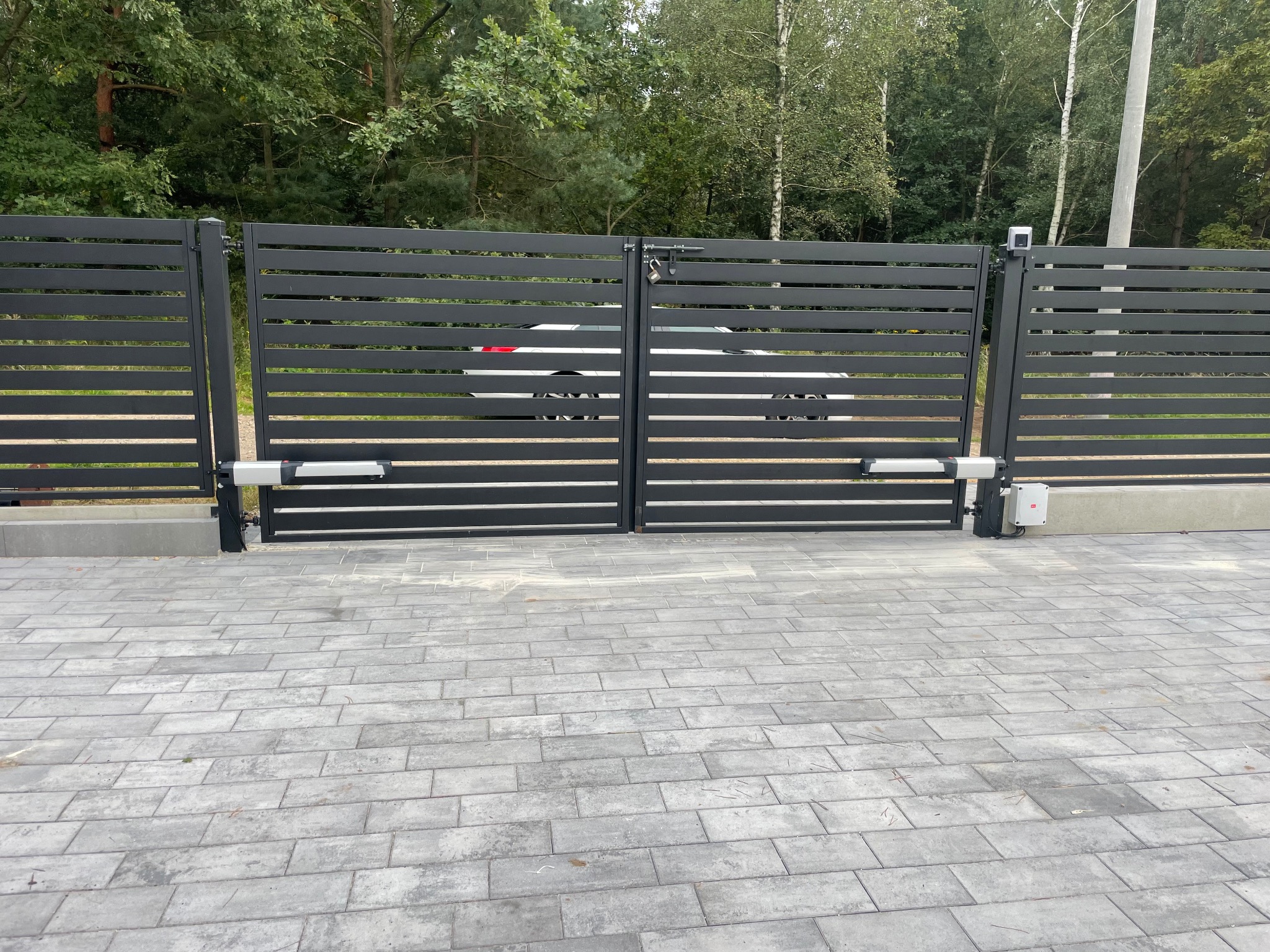 Nowoczesna, grafitowa brama przesuwna z automatyką, widoczna z zewnątrz posesji na tle zieleni. Kostka brukowa przed bramą.