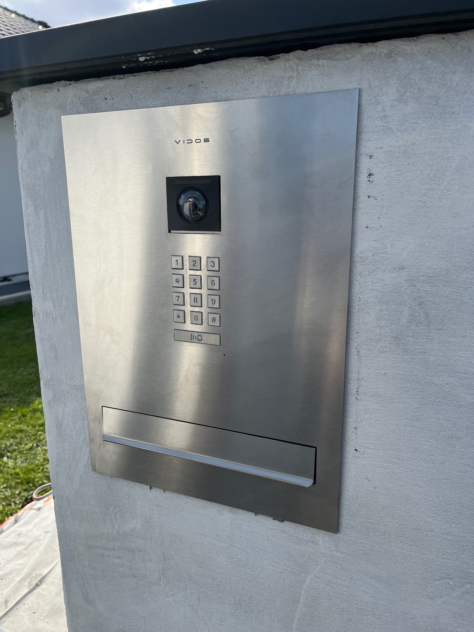 Nowoczesny, srebrny panel domofonowy Vidos z kamerą i klawiaturą numeryczną, zamontowany na szarym, surowym słupku ogrodzeniowym przy domu.