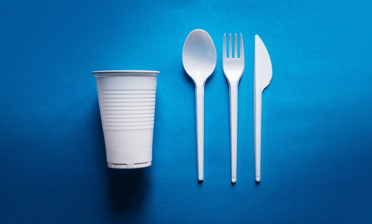 Biały plastikowy kubek, łyżka, widelec i nóż ułożone obok siebie na niebieskim tle, widok z góry.