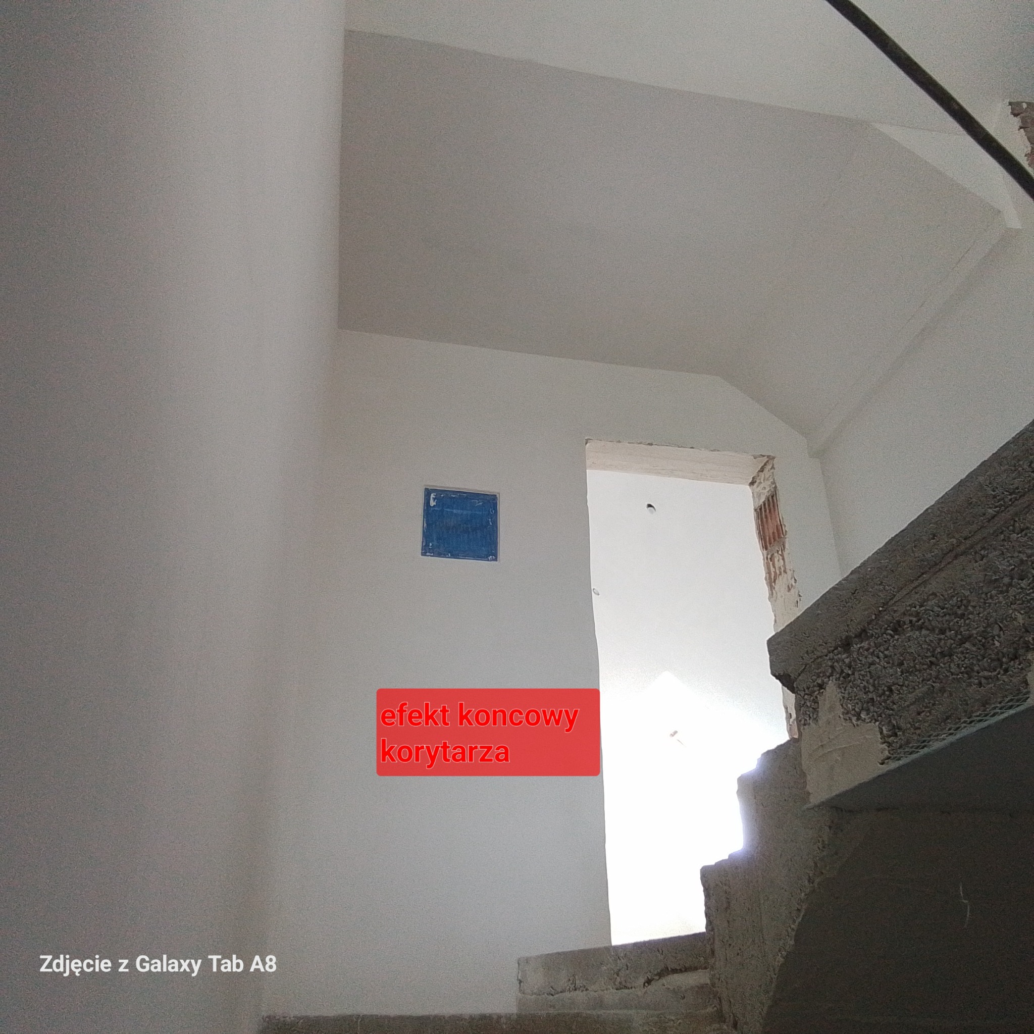 Korytarz po remoncie, świeżo pomalowane ściany na biało, widoczne schody w stanie surowym, zabezpieczone niebieską folią gniazdko elektryczne na ścianie.