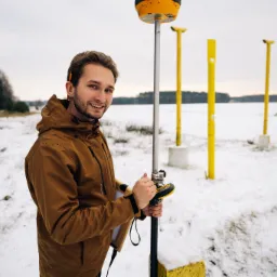 Młody mężczyzna w brązowej kurtce z kapturem, z uśmiechem, obsługuje żółty odbiornik GPS na tle zimowego krajobrazu z żółtymi słupkami.