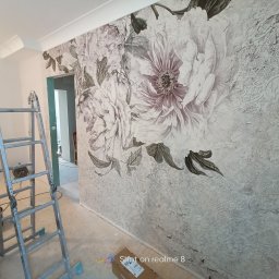 Marcin Polak - Ściana z dekoracyjną tapetą w kwiaty i ornamenty, drabina aluminiowa w tle, karton na podłodze.