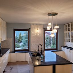 Marcin Polak - Wnętrze kuchni w trakcie prac wykończeniowych: białe szafki, czarny blat, mozaika herringbone na ścianie, wisząca lampa z kilkoma szklanymi kloszami, okna z widokiem na zieleń, podłoga...