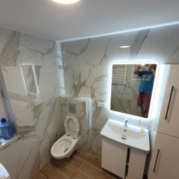 Marcin Polak - Wykończona łazienka z toaletą podwieszaną, umywalką z szafką, lustrem z podświetleniem LED i płytkami imitującymi marmur, widoczne środki czystości na parapecie okna.