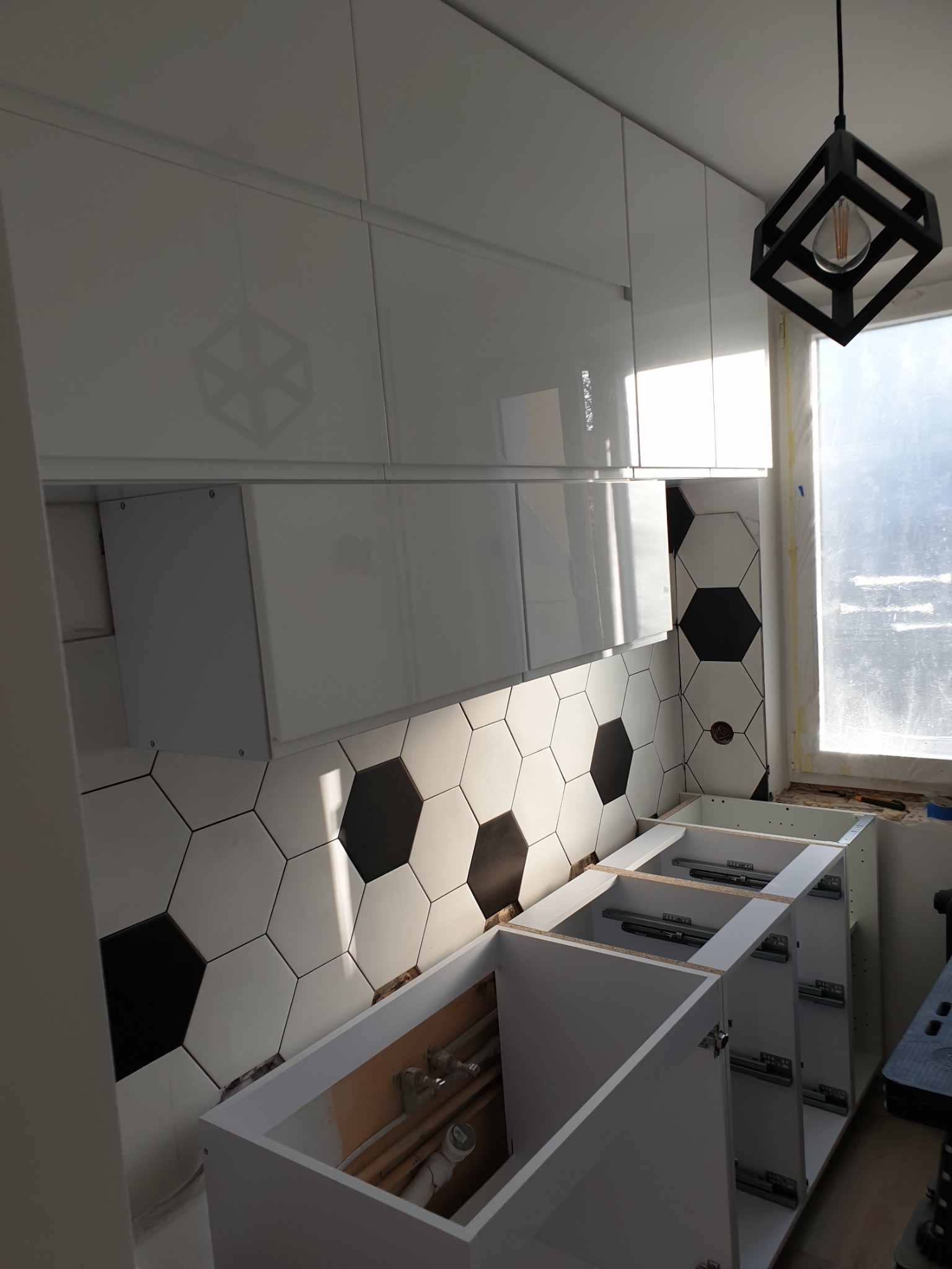 Montaż szafek kuchennych z czarno-białymi heksagonalnymi płytkami na ścianie i nowoczesną lampą wiszącą w kształcie sześcianu.