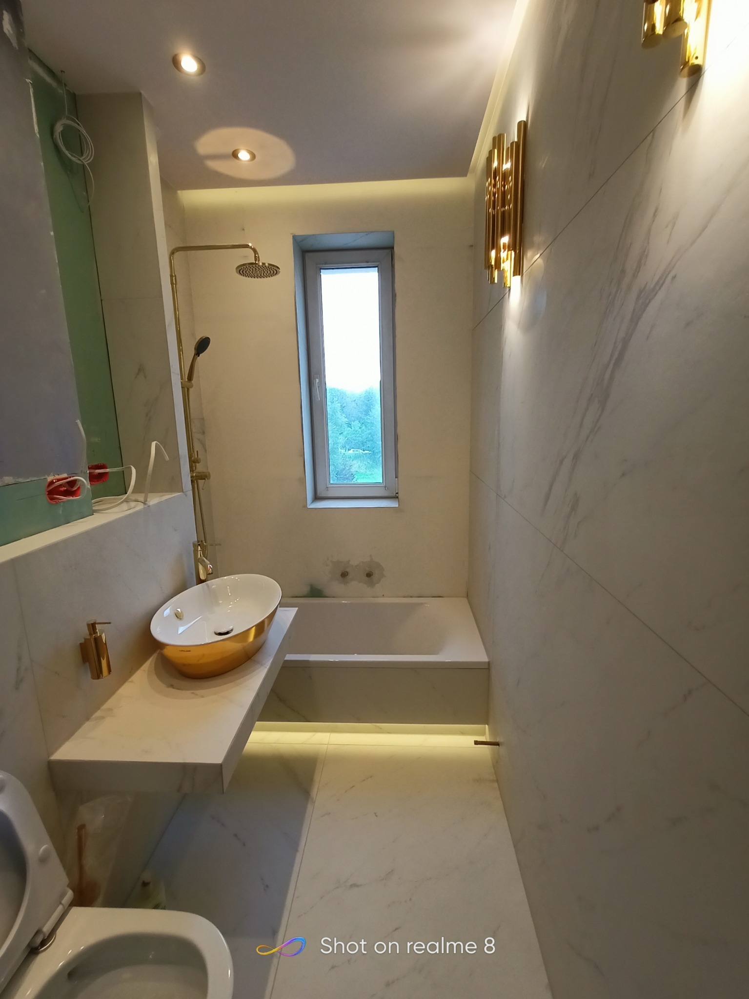 Nowoczesna łazienka w trakcie wykańczania, widoczna wanna, umywalka w kolorze złota, armatura w kolorze złota, białe płytki imitujące marmur, oświetlenie LED i lampy ścienne w kolorze złota...