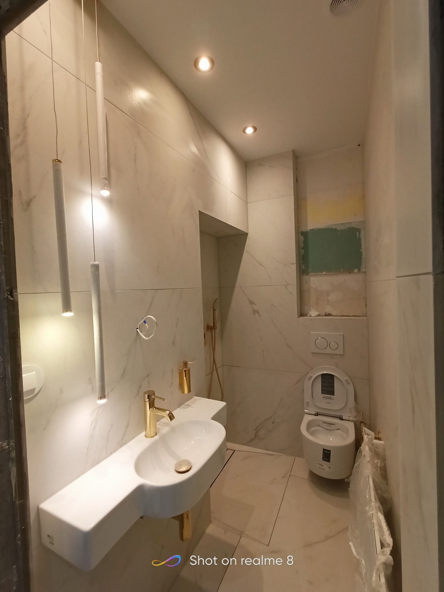Nowoczesna łazienka w trakcie wykańczania: białe marmurowe płytki, podwieszana umywalka z złotą baterią, wiszące lampy tubowe, toaleta w folii ochronnej, widoczne ślady prac malarskich.