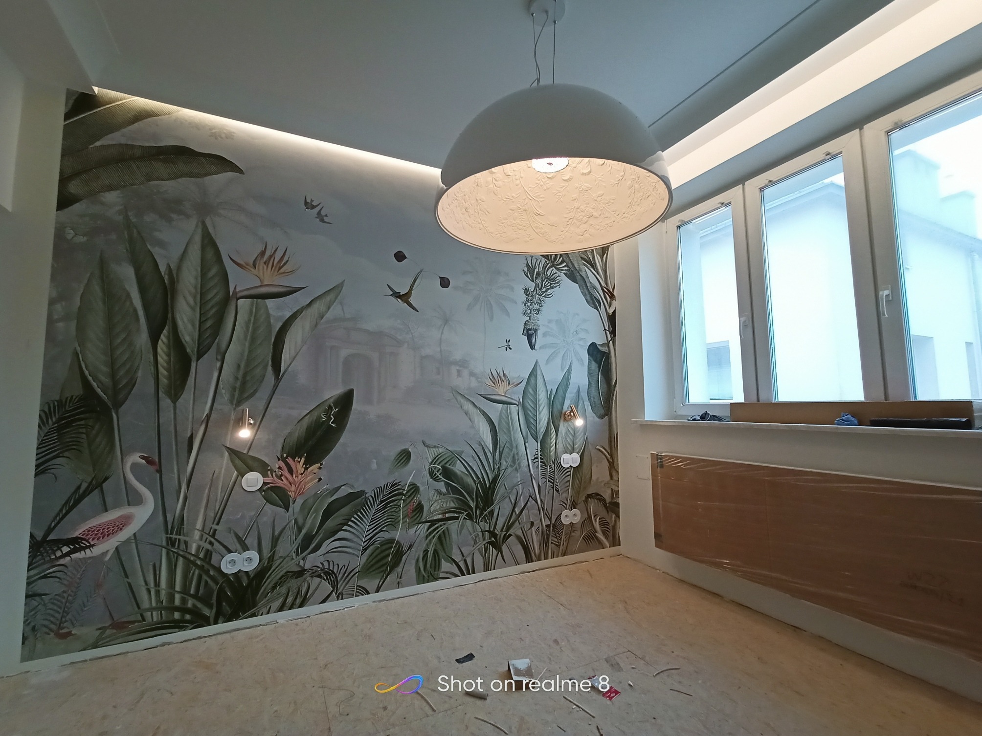 Pokój w trakcie wykańczania z fototapetą w motywy tropikalne, białą lampą wiszącą z ażurowym wzorem, widoczne gniazdka elektryczne i grzejnik zabezpieczony folią.